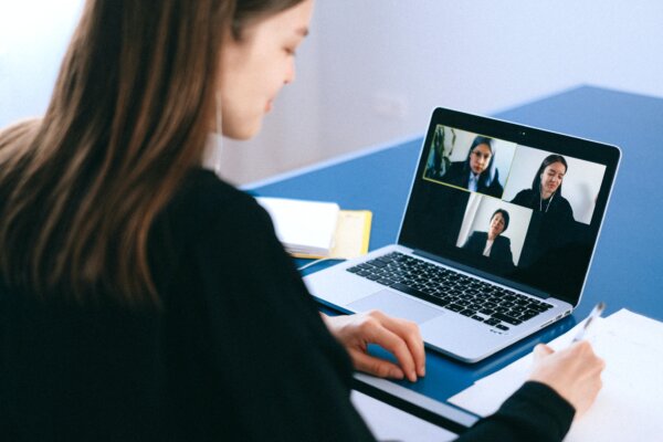 A woman sat at a desk with three other women on her laptop screen on an online meeting. Looks like hybrid teaching.