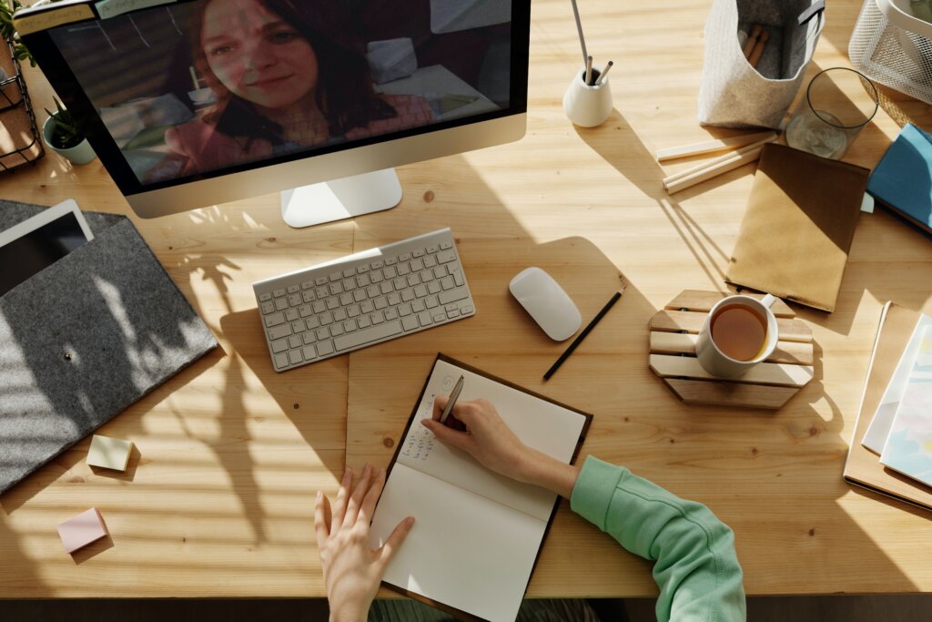 Person writing in notebook with a woman on the computer screen on an online meeting, possibly demonstrating hybrid teaching. Cluttered desk. 