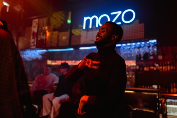 A man smiling and dancing with a bar and neon sign behind him