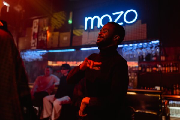 A man smiling and dancing with a bar and neon sign behind him