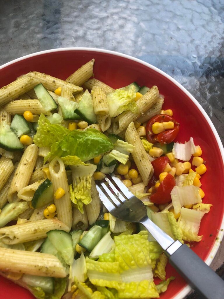 Pesto pasta in red bowl with fork and cherry tomatoes, tinned sweetcorn, cucumber chunks, lettuce 