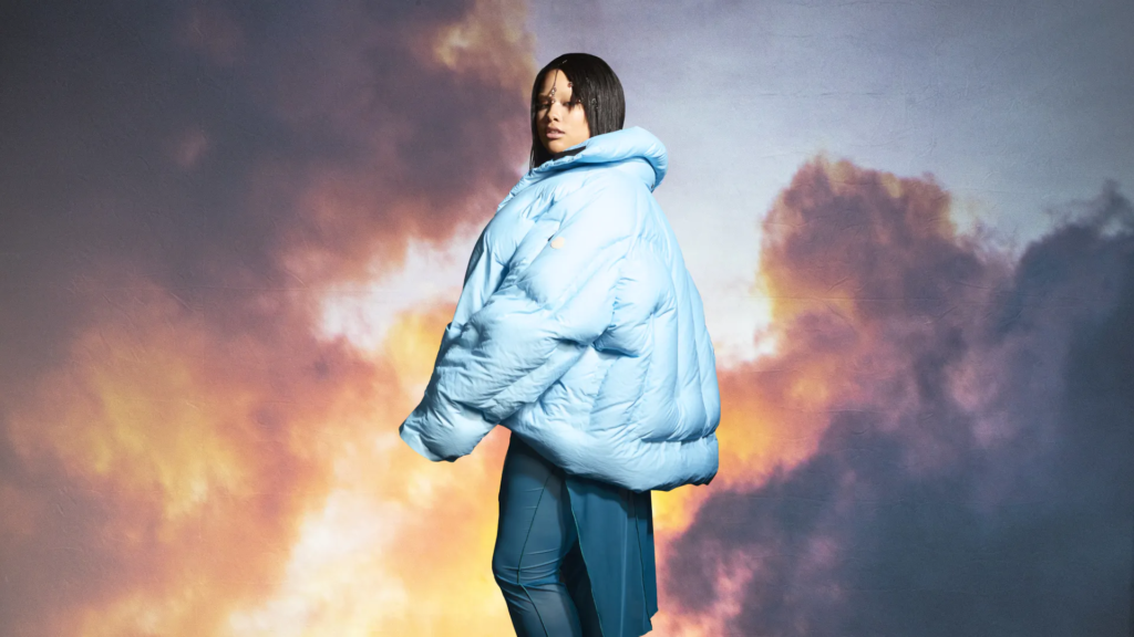 Picture of artist Shygirl in blue long puffer jacket. She is wearing dark blue trousers and standing against a wallpaper of a sky with orange and purple clouds.